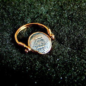 K&ouml;nnte beinhalten: Ein goldener Ring mit einer silbernen M&uuml;nze, die ein griechisches Design und die Inschrift "AVRELI" auf einem dunklen Hintergrund zeigt.