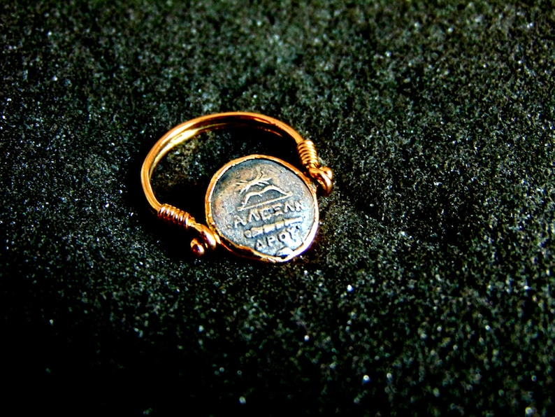 K&ouml;nnte beinhalten: Ein goldener Ring mit einer silbernen M&uuml;nze, die ein griechisches Design und die Inschrift "AVRELI" auf einem dunklen Hintergrund zeigt.