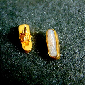 May include: Two gold-coloured rectangular stud earrings. One earring shows the back with a floral design and post. The other earring displays a light-coloured, oblong stone set in a gold frame. Jewellery.