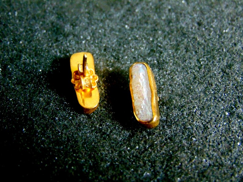 May include: Two gold-coloured rectangular stud earrings. One earring shows the back with a floral design and post. The other earring displays a light-coloured, oblong stone set in a gold frame. Jewellery.