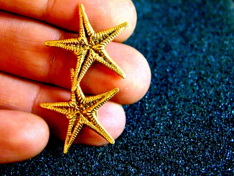 May include: Two gold-colored starfish charms, one on top of the other, with a textured surface. The charms are held in a hand.