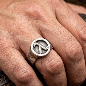 Op de afbeelding: Een zilverkleurige metalen ring met een cirkelvormig voorvlak en een verhoogd Grieks lambda-symbool. De ringband heeft een getextureerde, gearceerde afwerking die zich uitstrekt vanaf de cirkelvormige zetting langs de zijkanten van de band, wat zorgt voor een verweerde uitstraling.