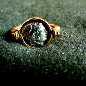 K&ouml;nnte beinhalten: Ein goldener Ring mit einer silbernen M&uuml;nze, die ein Profil eines menschlichen Kopfes zeigt. Die M&uuml;nze ist in einer Zargenfassung gefasst und der Ring ist verstellbar.