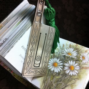 May include: A silver bookmark with a green tassel and a decorative design. The bookmark is on top of a book with the text "EUTUXIA" and "JEANET 15 A" visible.