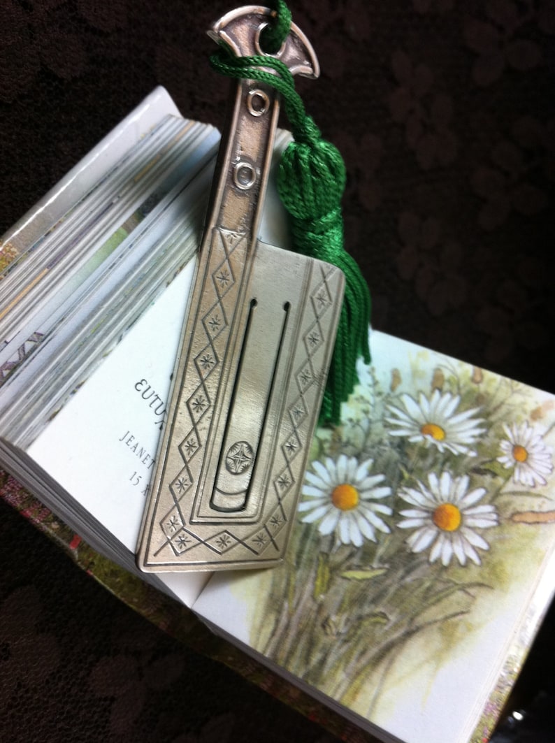 May include: A silver bookmark with a green tassel and a decorative design. The bookmark is on top of a book with the text "EUTUXIA" and "JEANET 15 A" visible.