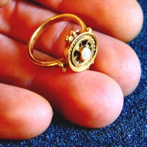 May include: A gold ring with an ornate design. The ring features a central, circular setting with a small, white gemstone. The band and setting are a bright gold color. The ring is held in a person's hand.