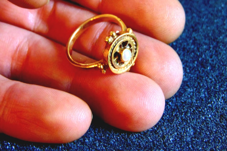 May include: A gold ring with an ornate design. The ring features a central, circular setting with a small, white gemstone. The band and setting are a bright gold color. The ring is held in a person's hand.