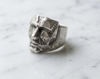 May include: A silver-toned metal ring featuring a sculpted human face with a textured, weathered finish. The design displays geometric, angular facial features including a prominent nose, closed eyes, and a defined mouth, all integrated into the band.