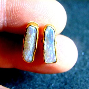 May include: A pair of gold-plated stud earrings. Each earring has a rectangular, iridescent, light-coloured stone with a gold border. The earrings are shown against a blurred background.