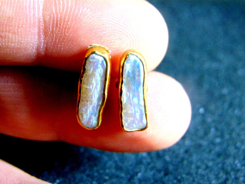 May include: A pair of gold-plated stud earrings. Each earring has a rectangular, iridescent, light-coloured stone with a gold border. The earrings are shown against a blurred background.