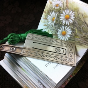 May include: A silver bookmark with a green tassel and a geometric design. The bookmark is resting on a book with a floral illustration and the text "EUTUX JEANNE 15".