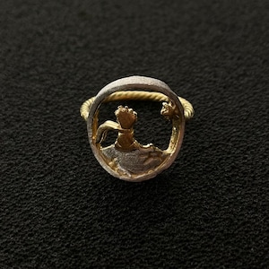 Puede incluir: Un anillo de plata con un diseño dorado de una persona que alcanza una rama de árbol. El anillo está sobre un fondo negro.