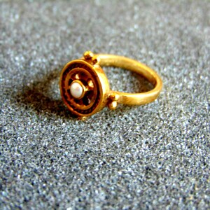 May include: A gold-colored ring with a circular design and a small, white pearl in the center. The ring features a detailed pattern around the pearl and on the sides. The ring is set against a textured, gray background.