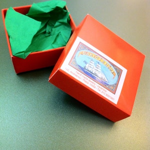 May include: A red box with a green tissue paper lining. The box has a label with the text "Gioielli di Abo" and the address "142, Andrianou - Plaka 10558 - Tel. 210 3231321". The label also features a hand-drawn illustration of a ship.
