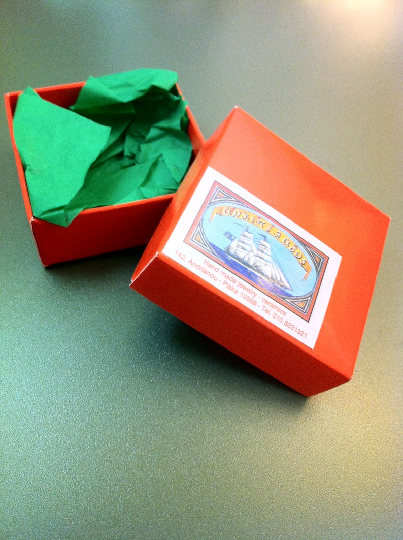 May include: A red box with a green tissue paper lining. The box has a label with the text "Gioielli di Abo" and the address "142, Andrianou - Plaka 10558 - Tel. 210 3231321". The label also features a hand-drawn illustration of a ship.