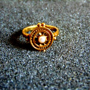 May include: A gold-colored ring with a circular design. The ring features a central pearl and an outer decorative pattern. The ring is set against a textured, dark gray surface.