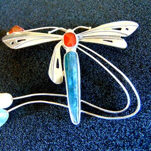 K&ouml;nnte beinhalten: Eine silberne Libellenbrosche mit blauen und roten Edelsteinen. Die Fl&uuml;gel der Libelle sind mit Ausschnitten verziert und der K&ouml;rper ist mit einem langen, blauen Edelstein geschm&uuml;ckt. Die Brosche hat ein zartes, flie&szlig;endes Design mit einer Perle und einem kleineren blauen Edelstein, der unten h&auml;ngt.
