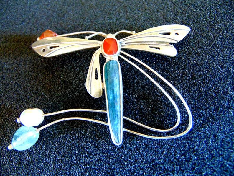 K&ouml;nnte beinhalten: Eine silberne Libellenbrosche mit blauen und roten Edelsteinen. Die Fl&uuml;gel der Libelle sind mit Ausschnitten verziert und der K&ouml;rper ist mit einem langen, blauen Edelstein geschm&uuml;ckt. Die Brosche hat ein zartes, flie&szlig;endes Design mit einer Perle und einem kleineren blauen Edelstein, der unten h&auml;ngt.