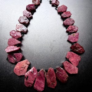 May include: A necklace strand featuring raw, irregularly shaped ruby gemstones in shades of deep pink and magenta. The stones are strung together with small, round, black spacer beads on a thin wire, creating a continuous circular arrangement.