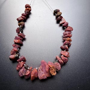 May include: A strand of raw, unpolished ruby gemstones in varying shades of deep pink, magenta, and dark red. The stones are irregularly shaped, rough-cut, and strung together on a thin wire with small white spacer beads between each gemstone.