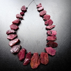 Peut inclure: Un collier composé de pierres précieuses rubis brutes, de forme irrégulière et de couleur rose foncé. Les pierres sont intercalées avec de petites perles d'espacement noires rondes et enfilées sur un fil fin et flexible visible en haut.