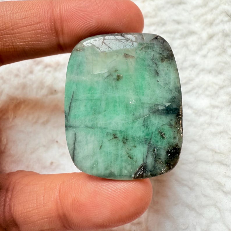 May include: A rectangular emerald gemstone with rounded corners. The stone has a vibrant green hue with darker green and black inclusions. The surface reflects light, showcasing natural patterns and textures. The gemstone is held between fingers.