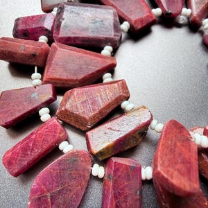 May include: A strand of raw, deep red ruby gemstones with irregular, faceted shapes. The stones are strung together with small, opaque white spacer beads, creating a textured, multi-piece necklace or jewelry component with a natural finish.