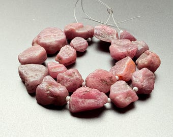 May include: A strand of rough-cut, pink gemstones. The stones vary in size and shape, strung together with a thin, white cord. The gemstones have a natural, unpolished appearance, with a range of pink hues. The strand is displayed against a gray background.