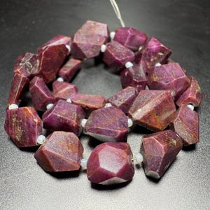 May include: Strung faceted ruby gemstone beads. The beads are a deep reddish-purple color with a faceted, irregular shape. Small white beads separate the ruby stones. The beads are strung on a white string.