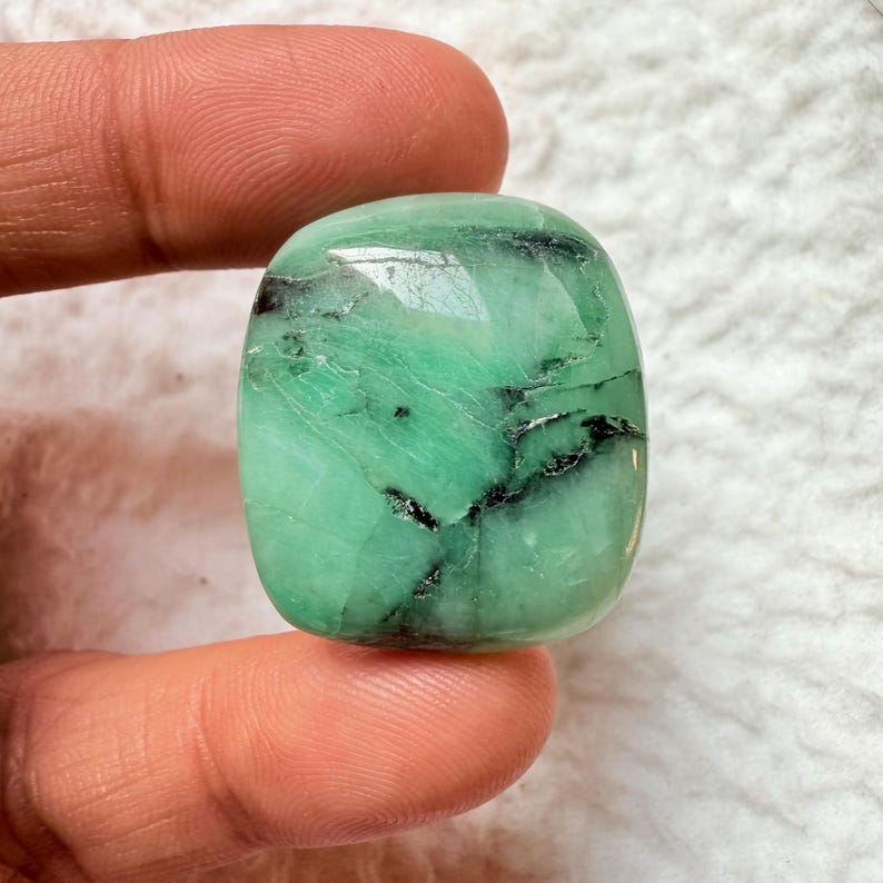 May include: A polished, emerald-green gemstone with black inclusions. The stone has a rounded, rectangular shape and a glossy surface. The gem is held between two fingers, showcasing its color and texture. The gemstone is a natural mineral.