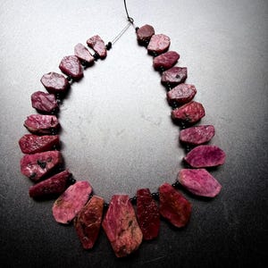 Pode incluir: Um colar composto por pedras preciosas de rubi brutas e de formato irregular em tons profundos de magenta e carmesim. As pedras são intercaladas com pequenas contas pretas redondas e enfiadas num cordão fino e escuro em disposição graduada.