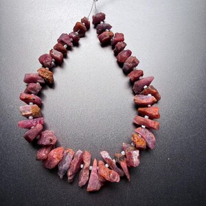 May include: A strand of raw, unpolished ruby gemstone beads arranged in a teardrop shape. The stones feature irregular, jagged edges and varying shades of deep red, pink, and brownish-orange, separated by small, round, white spacer beads on a string.