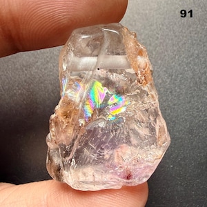 Puede incluir: Un cristal en bruto transparente con iridiscencia de arcoíris. El cristal tiene una textura rugosa y natural y una mezcla de tonos claros y marrones. El número "91" es visible en la esquina superior derecha. Este cristal es una gema natural única.