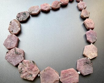 May include: A necklace featuring rough-cut, hexagonal pink gemstones strung on a wire. The stones have a natural, unpolished appearance with variations in color and texture, and are connected by small white beads.