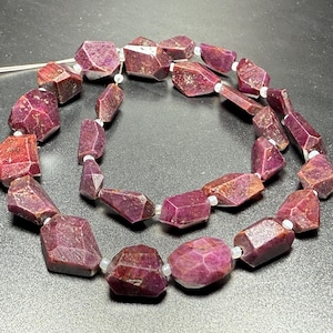 May include: Strand of faceted ruby beads. The beads are a deep red-purple colour with variations in tone. Each bead is irregularly shaped and strung on a white cord, creating a necklace or bracelet.