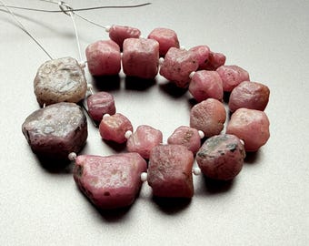 May include: A collection of rough-cut, pink gemstone beads strung on thin wire. The beads vary in size and shape, with a natural, unpolished appearance. The color ranges from light to deep pink, with some beads showing darker inclusions. The beads are ready for jewelry making.