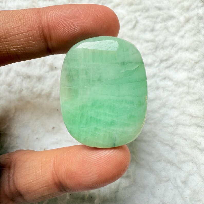 May include: A polished, oval-shaped gemstone in shades of light green. The stone has a smooth, glossy surface with visible internal patterns. The gemstone is held between two fingers, showcasing its size and color.