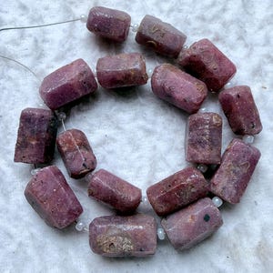 Puede incluir: Un hilo de cuentas de rubí en bruto de forma cilíndrica en tonos variados de rosa intenso y magenta. Cada cuenta presenta una textura natural rugosa con inclusiones oscuras visibles y superficies cristalinas, ensartadas en un alambre fino y flexible.