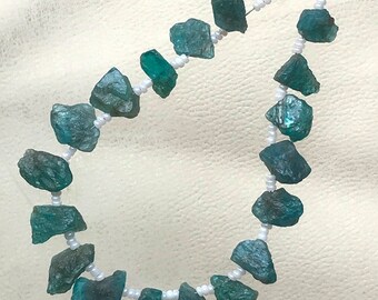 Puede incluir: Un collar con piedras preciosas de color turquesa en bruto, ensartadas en un hilo de cuentas blancas. Las piedras tienen una forma irregular y están espaciadas a lo largo del hilo. El fondo es una tela blanca suave.