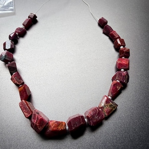 May include: A strand of raw, deep red ruby gemstone beads, each featuring an irregular, faceted, and chunky shape. The stones are strung on a thin, silver-colored wire, with small, white, round spacer beads placed between each of the larger red gems.