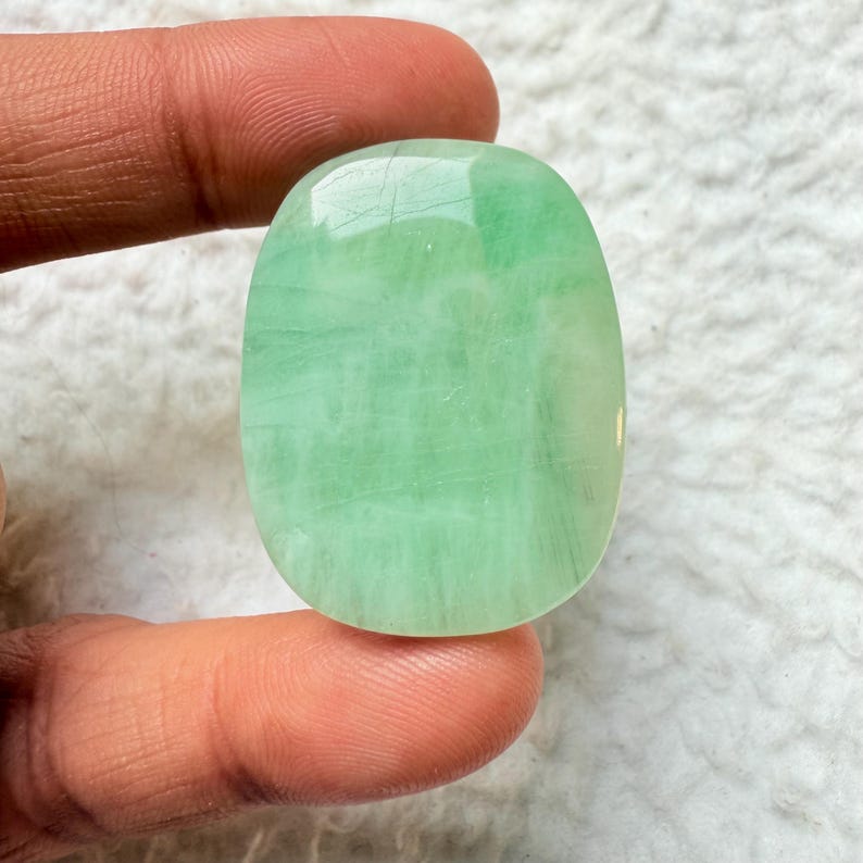 May include: A polished, oval-shaped gemstone with a translucent, pale green hue. The stone displays subtle internal patterns and a smooth, glossy surface. The gemstone is held between two fingers.