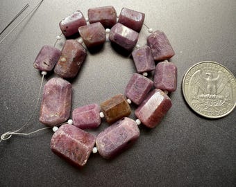May include: A strand of faceted, reddish-purple gemstone beads. The beads are rectangular with rounded edges and are strung on a white cord. A U.S. quarter coin is next to the beads for size comparison. The beads have a natural, earthy appearance.