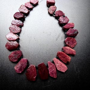 Peut inclure: Un collier composé d'un rang de rubis bruts, allant du rose profond au rouge foncé, aux formes irrégulières et facettées. Les pierres sont séparées par de petites perles noires rondes et enfilées sur un fil fin et sombre en cercle.