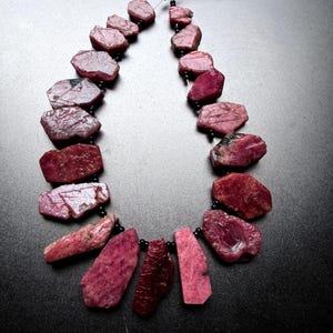 May include: A necklace strand featuring raw, irregularly shaped ruby gemstones in deep shades of magenta and crimson. The stones are interspersed with small, round, black spacer beads and are strung on a thin, metallic wire that remains open.