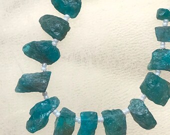 Puede incluir: Un collar con piedras preciosas en bruto de color turquesa, ensartadas con pequeñas cuentas blancas. Las piedras preciosas tienen una forma natural e irregular y diferentes tonos de turquesa. El collar se muestra sobre un fondo blanco suave.
