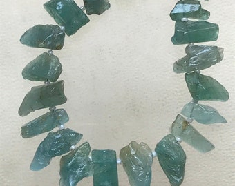 May include: A necklace featuring rough-cut, aquamarine-colored gemstones strung together with small white beads. The gemstones have irregular shapes and varying shades of blue-green. The necklace is displayed against a neutral background.