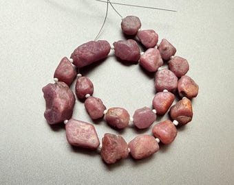 May include: A strand of rough-cut, pink-red gemstone beads. The beads are irregular in shape and size, strung together with white string. The gemstones have a natural, unpolished appearance. Ideal for jewelry making.