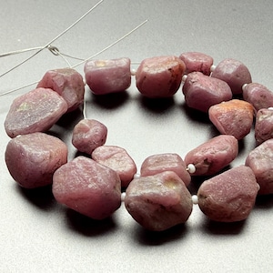 May include: A necklace of raw, pink-red ruby beads. The beads are of varying sizes and shapes, threaded on a fine wire. The gemstones have a natural, unpolished look, with differences in colour and texture.