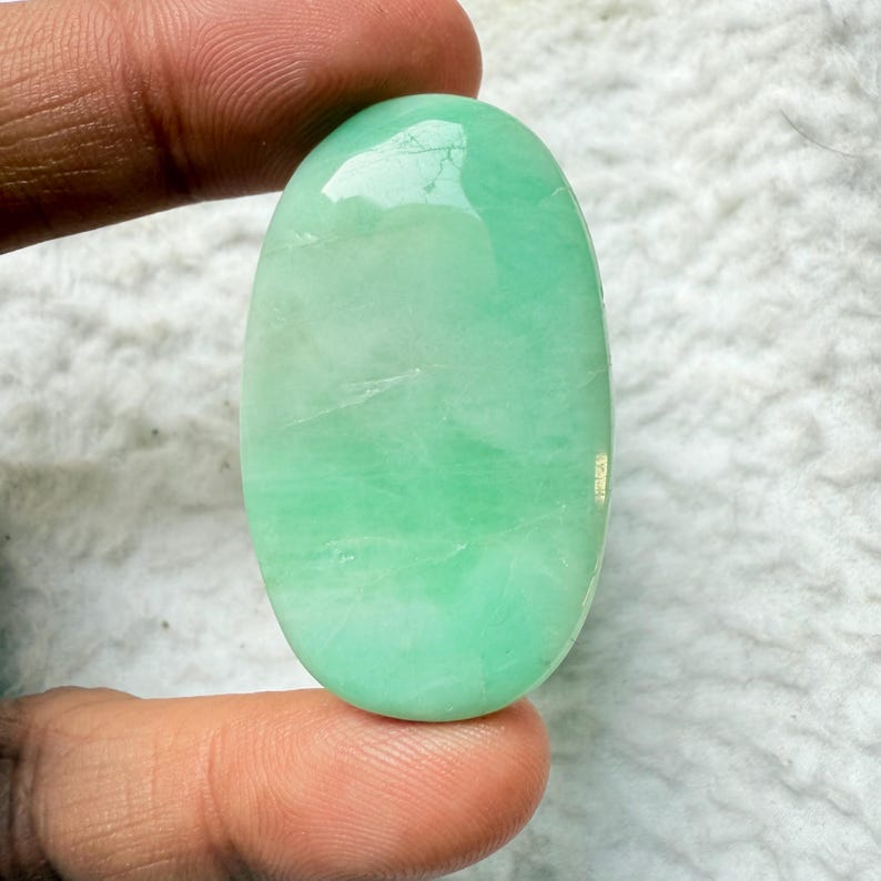 May include: A polished, oval-shaped gemstone in shades of light green. The stone has a smooth, reflective surface and appears to be held by a person's fingers. The gemstone's color is consistent throughout, with subtle variations in tone.