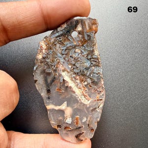 May include: A translucent, irregularly shaped stone with a drilled hole. The stone features a mix of brown, black, and blue patterns, with a white vein running through it. The number "69" is visible in the upper right corner.